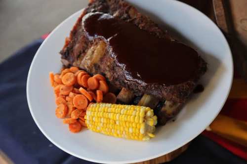 A plate featuring a meatloaf with barbecue sauce, served with corn on the cob and a side of sliced carrots.