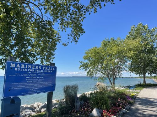 A scenic view of a waterfront trail with a sign about trail rules, surrounded by trees and colorful flowers.