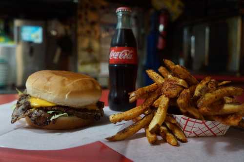 A cheeseburger with a soft bun, a bottle of Coca-Cola, and a side of seasoned fries on a red table.