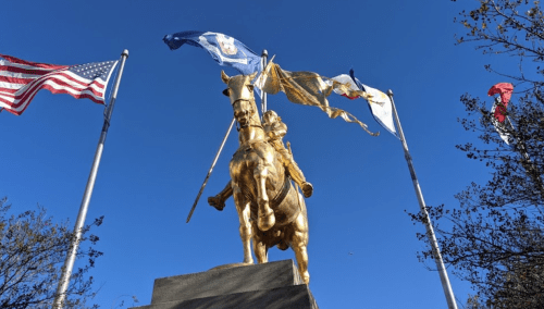 A golden statue of a mounted figure holding flags, surrounded by American and state flags against a clear blue sky.