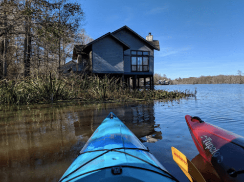 A view from a kayak on calm water, featuring a blue house on stilts surrounded by trees and reeds.