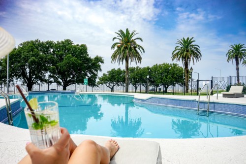 A person relaxes by a pool, holding a drink, with palm trees and a clear sky in the background.