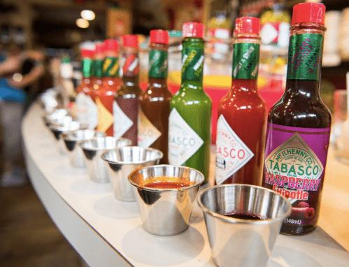 A row of various Tabasco sauce bottles with small metal tasting cups in front, displayed in a store.