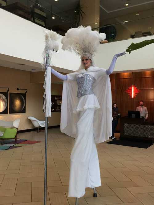 A performer on stilts in a white costume with feathers, standing in a hotel lobby with guests in the background.