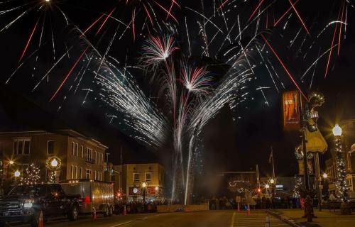Colorful fireworks burst in the night sky over a festive street, with buildings and a crowd visible below.