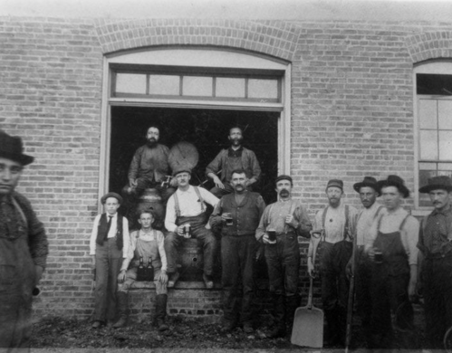 Black and white photo of a group of workers posing outside a brick building, some holding tools and drinks.