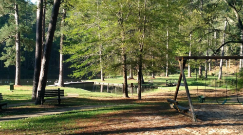 A serene park scene with a swing set, benches, and a calm lake surrounded by trees.