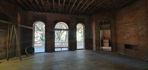 Interior of a brick room with arched windows, wooden beams, and a wooden floor, featuring a view of trees outside.