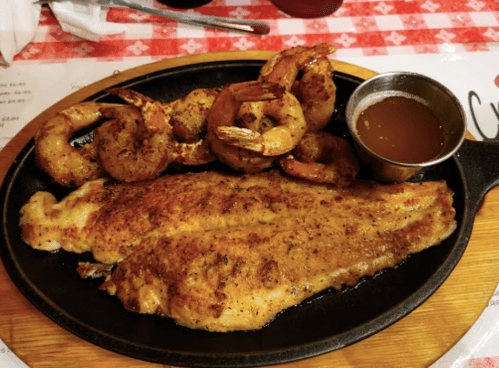 A sizzling platter with seasoned fish fillet, grilled shrimp, and a side of dipping sauce on a checkered tablecloth.