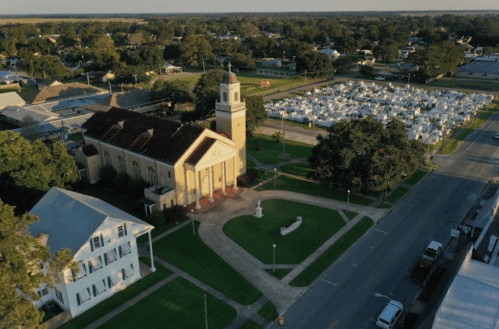 Aerial view of a church with a clock tower, surrounded by green lawns and a cemetery in the background.