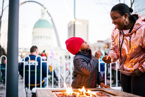 A woman and child enjoy roasting marshmallows by a fire pit, with a city skyline and arch in the background.