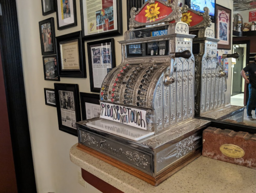 A vintage cash register on a countertop, with a sign reading "Please Do Not Touch," surrounded by framed photos on the wall.