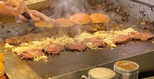A grill with sizzling patties and onions, with burger buns in the background, creating a smoky cooking scene.