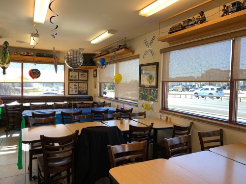 A cozy restaurant interior with tables, balloons, and decorations, featuring large windows and a view of the street outside.
