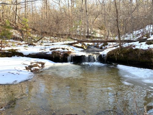 A serene winter scene featuring a small waterfall flowing into a calm stream, surrounded by snow and bare trees.