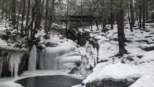 A snowy landscape featuring a frozen waterfall, icy rocks, and a bridge in the background surrounded by trees.