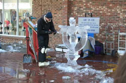 A sculptor carves an ice statue in a brick-paved area, surrounded by ice shavings and tools.