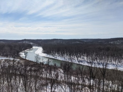 A winding river flows through a snowy landscape, surrounded by bare trees and rolling hills under a cloudy sky.