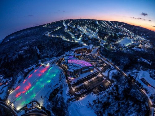 Aerial view of a snowy landscape with illuminated ski slopes and a resort at dusk, showcasing vibrant colors and winter scenery.