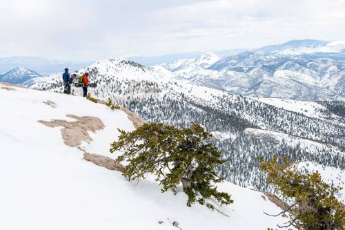 Two hikers stand on a snowy mountain ridge, overlooking a vast landscape of snow-covered peaks and valleys.