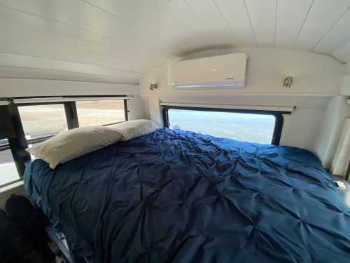 Cozy bedroom with a blue comforter and two pillows, featuring windows and an air conditioning unit above.