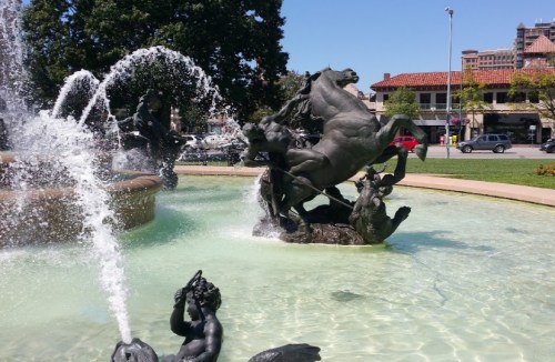 A decorative fountain featuring bronze sculptures of horses and figures, surrounded by clear water and greenery.