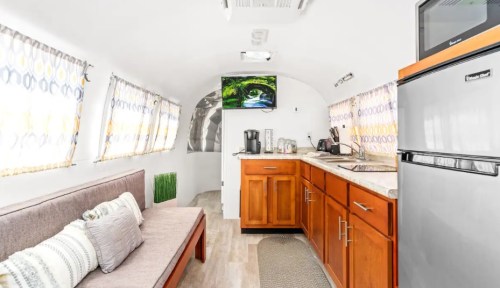 Interior of a modern, compact trailer featuring a sofa, kitchenette, and a small TV, with bright curtains and wood accents.