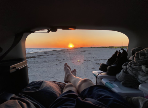 A person relaxes in a car, watching a sunset over a beach with bare feet visible and a serene atmosphere.