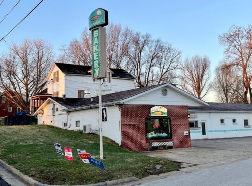 A small bakery with a sign, located on a grassy hill, surrounded by trees and a few for-sale signs.