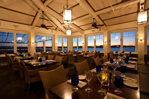 A beautifully set dining room with blue glassware, warm lighting, and large windows showcasing a sunset over water.