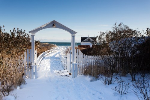 A snowy pathway leads through a white archway to a beach house by the ocean, framed by shrubs and a clear blue sky.