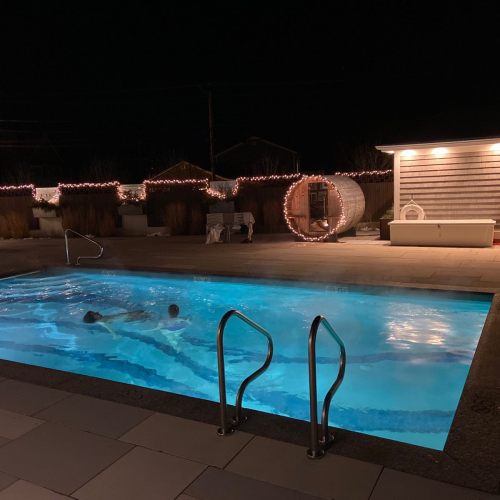 A person swims in a lit pool at night, surrounded by festive lights and a cozy outdoor setting.