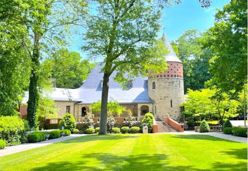 A picturesque stone castle surrounded by lush greenery and manicured lawns under a clear blue sky.