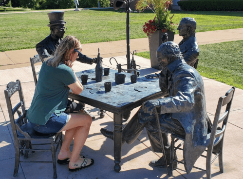 A woman sits at a table with bronze statues of four men, engaged in conversation in a park setting.