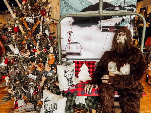 A cozy holiday scene featuring a Sasquatch figure sitting on a bench next to a decorated Christmas tree and ski lift backdrop.