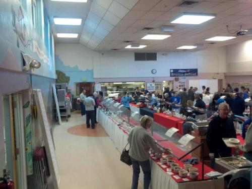 A busy community hall with people browsing a long buffet table filled with food and various dishes.