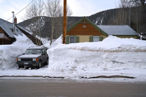 A house with a green and yellow facade, surrounded by tall snowbanks, and a parked car partially buried in snow.
