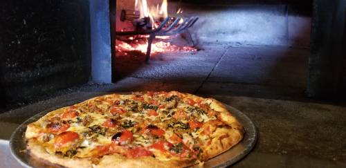 A freshly baked pizza with pepperoni and herbs, placed in front of a wood-fired oven with flames visible.