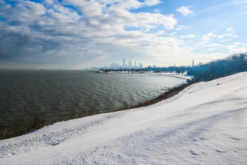A snowy shoreline with a view of a city skyline under a cloudy sky, reflecting on the water.