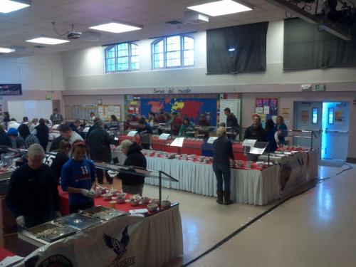 A busy cafeteria with people serving and enjoying food at multiple buffet stations, decorated with red tablecloths.