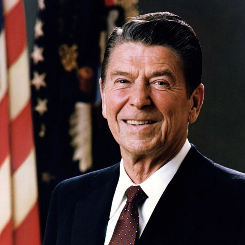 A smiling man in a suit with a tie, standing in front of an American flag.
