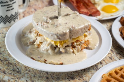 A plate of waffles topped with eggs and smothered in creamy sausage gravy, served with bacon and eggs in the background.