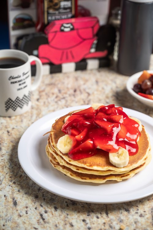 A stack of pancakes topped with banana slices and strawberry sauce, served with a cup of coffee in the background.