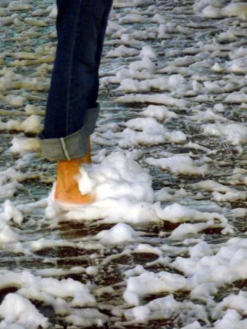 A person stands in shallow water covered with white foam, with rolled-up jeans and one foot submerged in the foam.