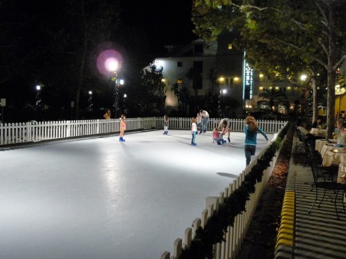 A nighttime ice skating rink with people skating, surrounded by trees and buildings, illuminated by lights.