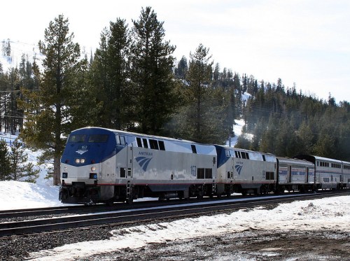 An Amtrak train travels through a snowy landscape, surrounded by evergreen trees and mountains in the background.