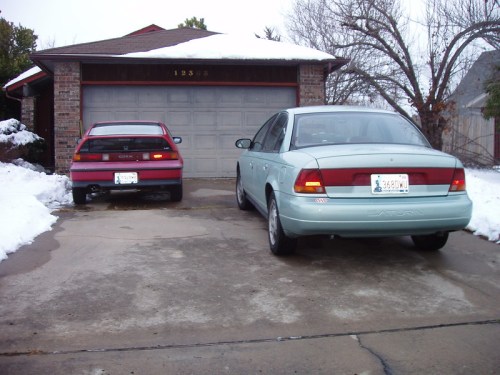 Two cars parked in a snowy driveway in front of a brick house with a garage door.