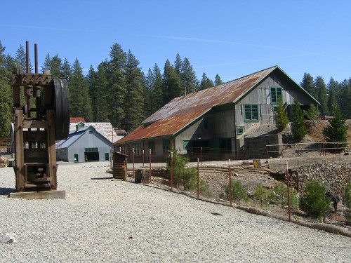 Abandoned mining buildings with a rusted structure in the foreground, surrounded by trees and a clear blue sky.