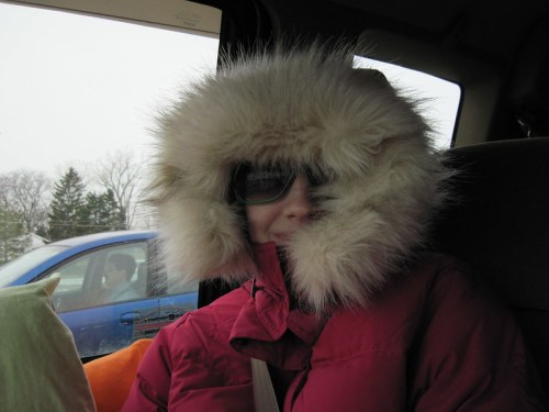 A person wearing a fur-lined hooded coat and sunglasses sits in a car, with a snowy landscape visible outside.