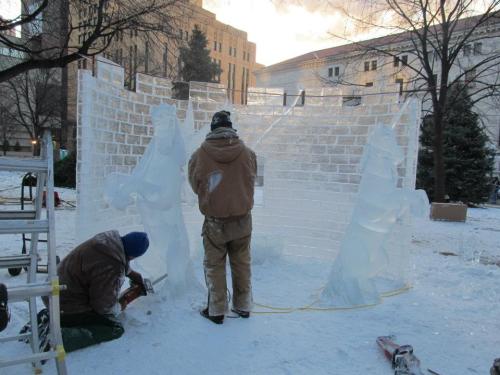 Two artists sculpt ice figures in a snowy outdoor setting, with a partially completed ice wall in the background.